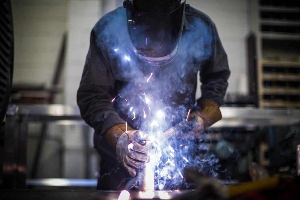  man, wearing protective gloves, jacket and mask, welds pieces of aluminum together with an arc welder.