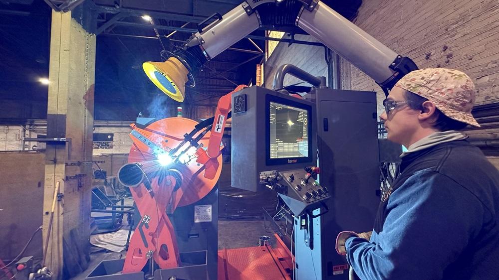 An operator stands at a machine as it welds a pipe. 