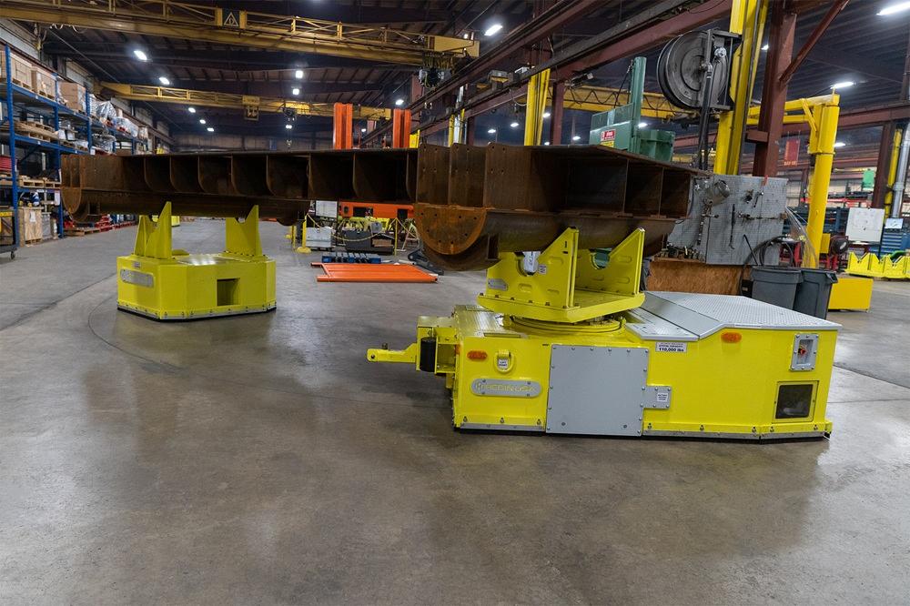 Two automated tuggers move a heavy workpiece along a shop floor.