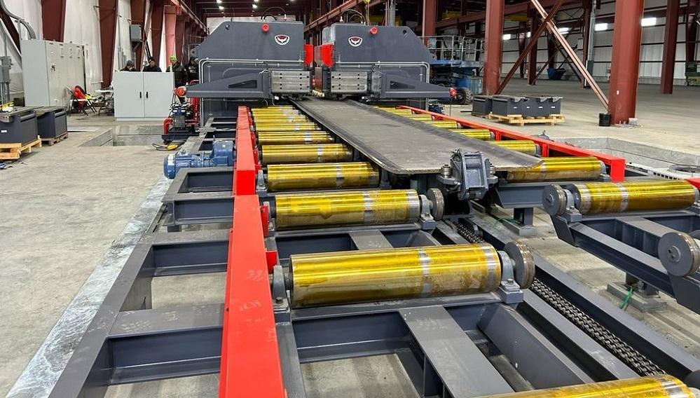 A long piece of metal sits on rollers in front of a bending machine.