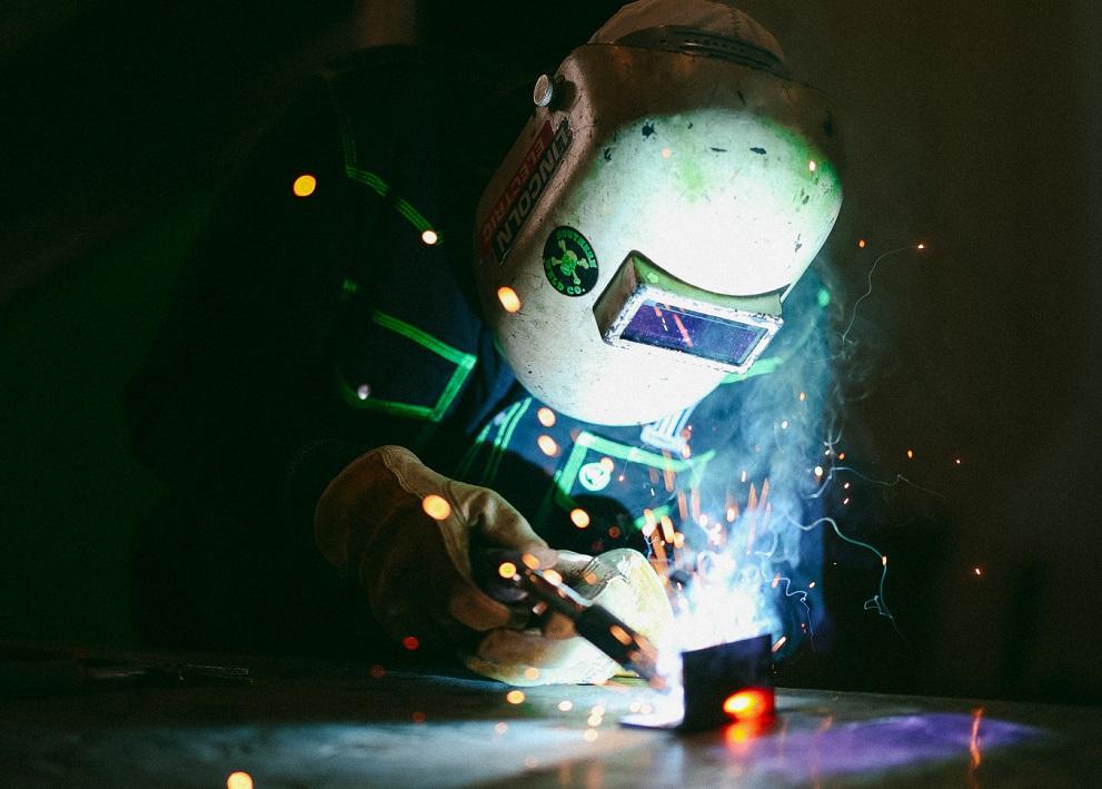 A person wearing a welding helmet welds a part.