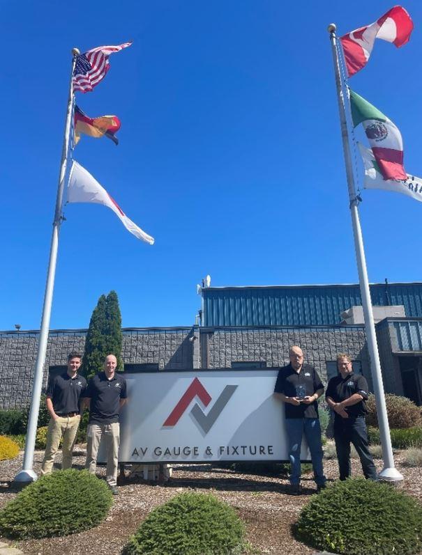 Four men stand in front of a company sign and flags.
