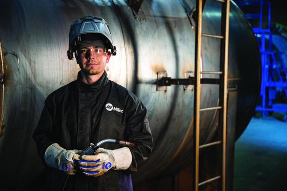 A welder stands in front of a metal tank. 