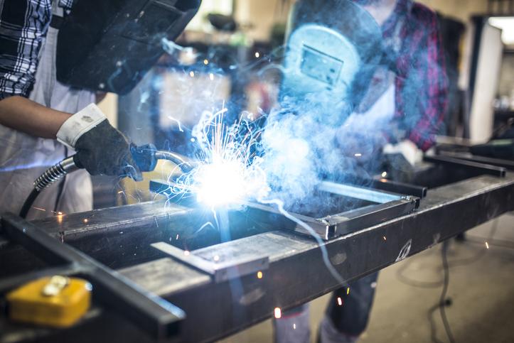 A young woman applies a MIG weld as an older man watches.