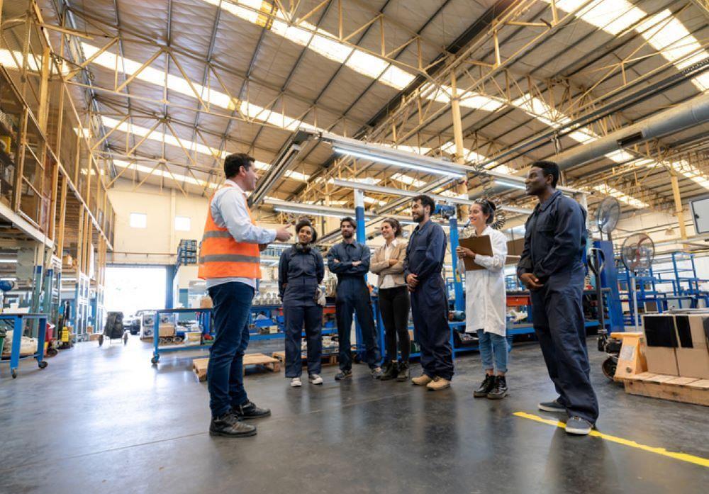 A manager instructs a diverse team of engineers, blue collar workers, and quality inspectors at a factory